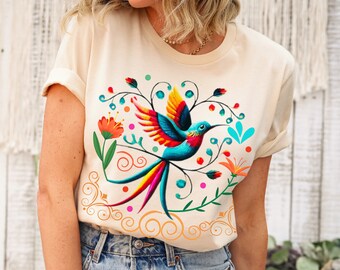 May include: A cream colored t-shirt with a colorful hummingbird and floral design. The hummingbird is blue, green, yellow, and red. The flowers are orange, red, and green.
