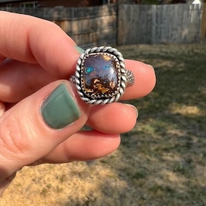 May include: A silver ring with a square, multi-colored opal stone. The opal features shades of brown, blue, and green. The ring has a twisted rope design around the stone and a textured band.