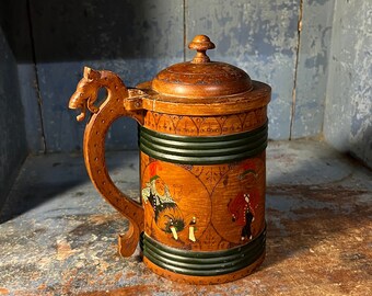 May include: A wooden beer stein with a carved handle in the shape of a dragon. The stein is decorated with green and black bands and painted figures in red, yellow, and blue.