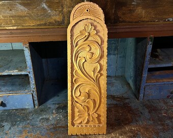 Puede incluir: Un colgante de pared de madera con intrincados diseños florales tallados. La pieza está fechada "1993" en la parte inferior.