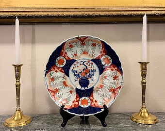 May include: A decorative plate with a blue, white, and orange floral design, displayed on a black stand. Two brass candlesticks with white candles flank the plate. A framed painting of a ship is in the background.