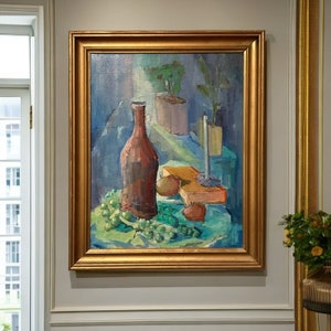 May include: A still life painting in a gold frame depicts a brown bottle, green grapes, a candle, and other objects on a blue tablecloth.
