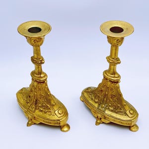 May include: Two ornate, gold-colored candle holders. Each has a round top with a central hole, a detailed stem, and a decorative base with small feet. The candle holders are made of metal and have an antique appearance.