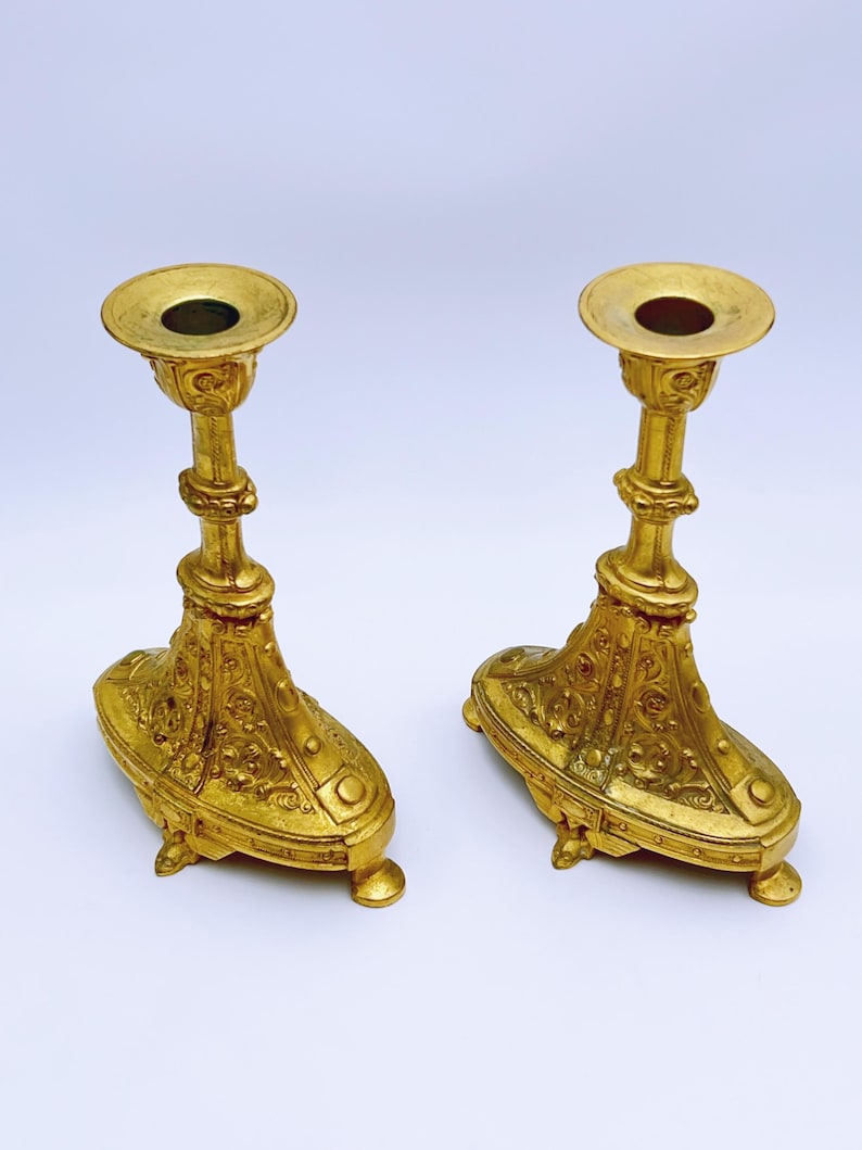 May include: Two ornate, gold-colored candle holders. Each has a round top with a central hole, a detailed stem, and a decorative base with small feet. The candle holders are made of metal and have an antique appearance.
