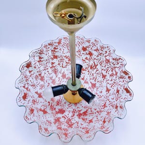 May include: A three-light ceiling fixture with a white glass shade featuring a red and white speckled pattern. The shade has a scalloped edge and is attached to a gold metal base.