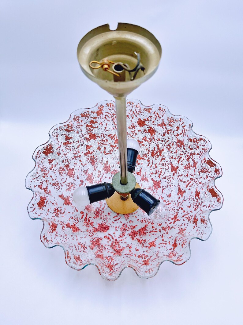 May include: A three-light ceiling fixture with a white glass shade featuring a red and white speckled pattern. The shade has a scalloped edge and is attached to a gold metal base.