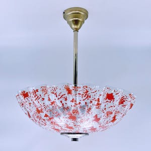 May include: A white ceiling light fixture with a red and white patterned glass shade. The shade has a scalloped edge and is suspended from a brass rod.