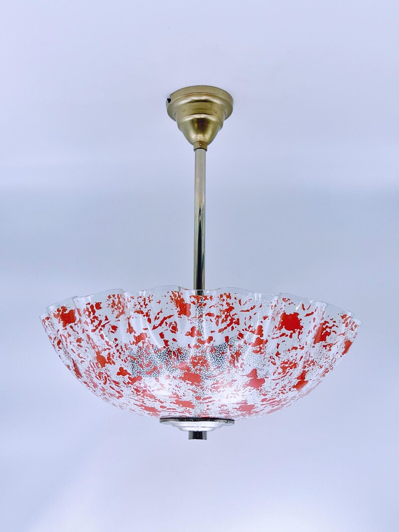 May include: A white ceiling light fixture with a red and white patterned glass shade. The shade has a scalloped edge and is suspended from a brass rod.