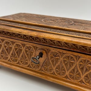 May include: A wooden box with intricate carved designs. The box is a warm brown color and features detailed patterns on the lid and front, including geometric shapes and floral motifs. A small key is in the keyhole.