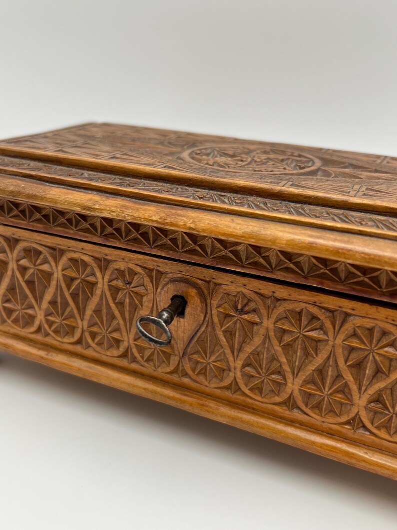 May include: A wooden box with intricate carved designs. The box is a warm brown color and features detailed patterns on the lid and front, including geometric shapes and floral motifs. A small key is in the keyhole.