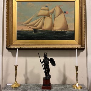 May include: A framed oil painting of a ship with white sails and the text "Jennie C" on the sail. The ship is sailing on a blue body of water with a small island in the background. The painting is hanging on a wall above a marble surface with two brass candle holders and a black metal statue of a warrior.
