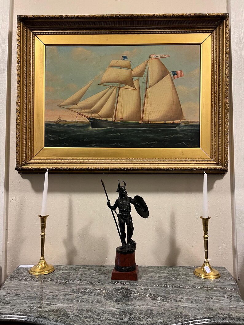 May include: A framed oil painting of a ship with white sails and the text "Jennie C" on the sail. The ship is sailing on a blue body of water with a small island in the background. The painting is hanging on a wall above a marble surface with two brass candle holders and a black metal statue of a warrior.