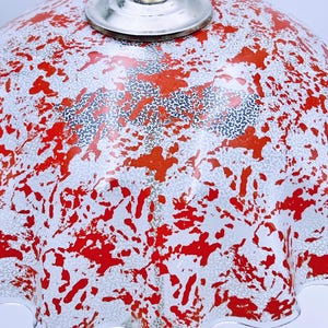 May include: A white lampshade with a red and silver splatter pattern. The lampshade has a ruffled edge and a silver metal top.