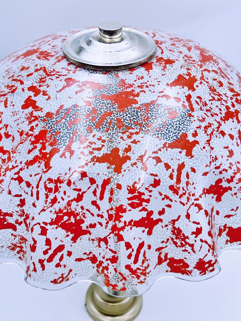 May include: A white lampshade with a red and silver splatter pattern. The lampshade has a ruffled edge and a silver metal top.