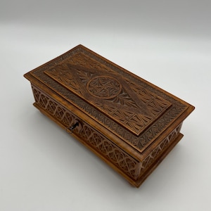 May include: A rectangular wooden box with intricate carved designs. The box is a warm brown color and features detailed geometric patterns on the lid and sides. A small keyhole is visible on the front.