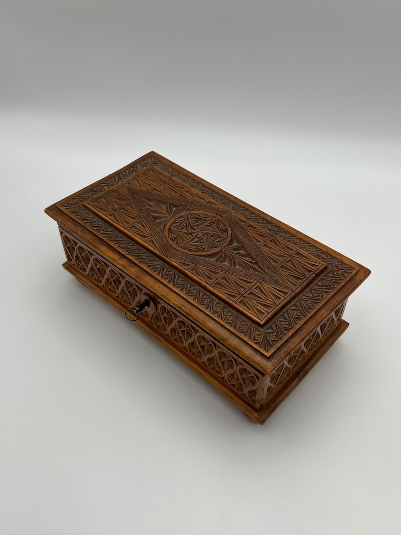 May include: A rectangular wooden box with intricate carved designs. The box is a warm brown color and features detailed geometric patterns on the lid and sides. A small keyhole is visible on the front.