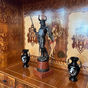May include: A black metal sculpture of a warrior with a spear and shield, standing on a red and black marble base. The sculpture is displayed on a wooden shelf with intricate marquetry designs.