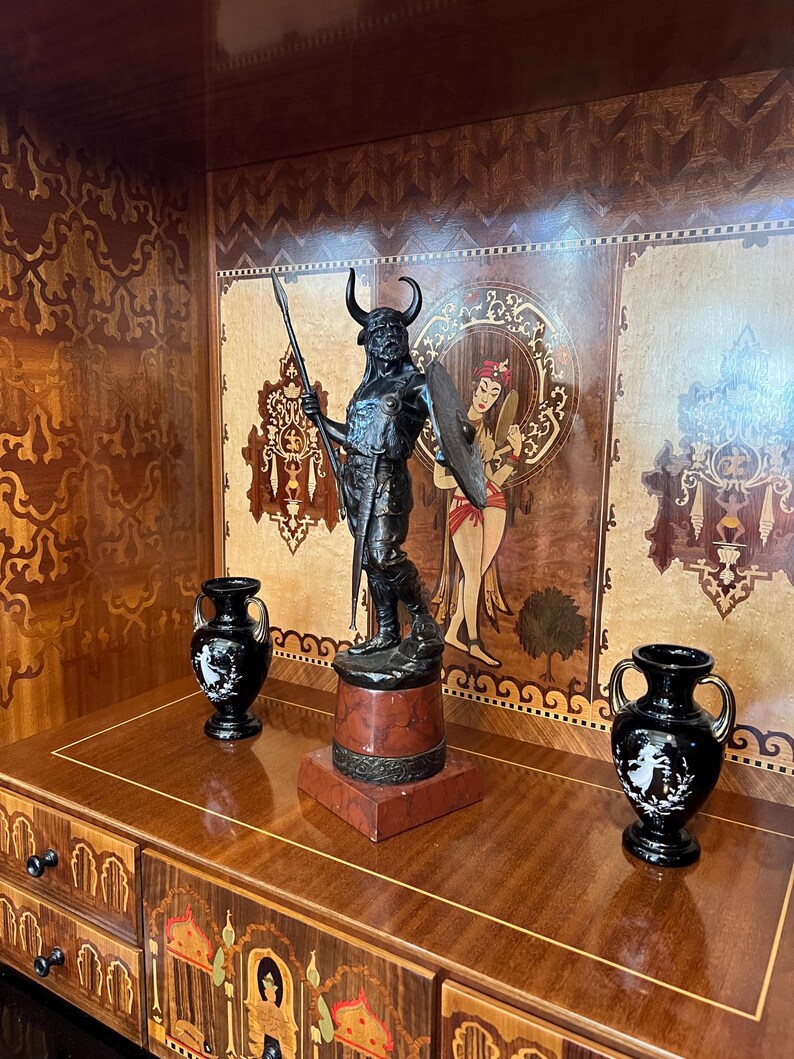 May include: A black metal sculpture of a warrior with a spear and shield, standing on a red and black marble base. The sculpture is displayed on a wooden shelf with intricate marquetry designs.