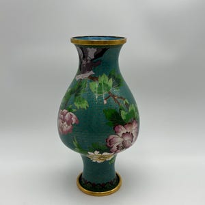 Puede incluir: Un jarr&oacute;n de cloisonn&eacute; turquesa con borde y base dorados. El jarr&oacute;n presenta un dise&ntilde;o floral con flores rosas y blancas, hojas verdes y un p&aacute;jaro. El fondo tiene un patr&oacute;n texturizado.