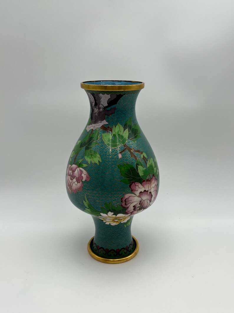 Puede incluir: Un jarr&oacute;n de cloisonn&eacute; turquesa con borde y base dorados. El jarr&oacute;n presenta un dise&ntilde;o floral con flores rosas y blancas, hojas verdes y un p&aacute;jaro. El fondo tiene un patr&oacute;n texturizado.