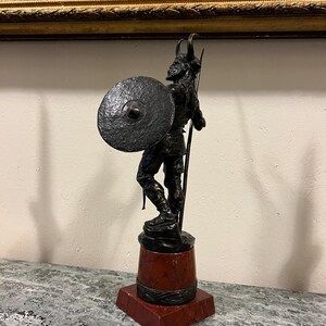 May include: A bronze sculpture of a Viking warrior holding a spear and shield. The sculpture is mounted on a red marble base.