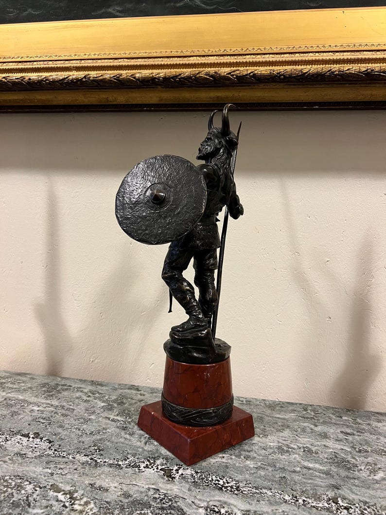 May include: A bronze sculpture of a Viking warrior holding a spear and shield. The sculpture is mounted on a red marble base.