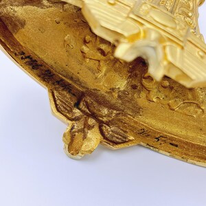 May include: Close-up of a gold-colored, ornate decorative object. The object has intricate details, including floral and geometric patterns. The base has a unique shape with a textured surface. The object appears to be made of metal.