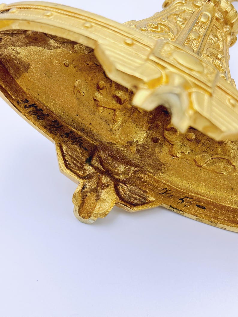 May include: Close-up of a gold-colored, ornate decorative object. The object has intricate details, including floral and geometric patterns. The base has a unique shape with a textured surface. The object appears to be made of metal.