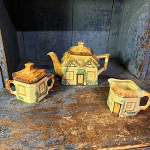 May include: A ceramic tea set featuring a teapot, sugar bowl, and creamer, all designed to resemble quaint cottages. The set has a yellow thatched roof and light green accents, with a door and windows on each piece. The set is displayed on a distressed wooden surface.