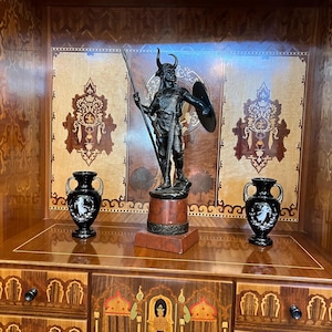 May include: A black metal statue of a warrior with a spear and shield, standing on a wooden base. The statue is displayed in a wooden cabinet with intricate marquetry details and two black ceramic vases.