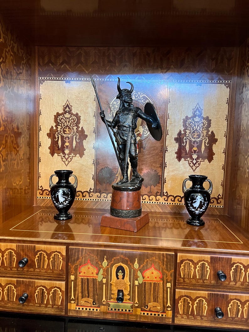 May include: A black metal statue of a warrior with a spear and shield, standing on a wooden base. The statue is displayed in a wooden cabinet with intricate marquetry details and two black ceramic vases.