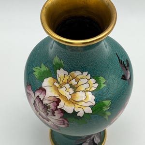 Puede incluir: Un jarr&oacute;n turquesa con borde y base dorados. El jarr&oacute;n presenta un dise&ntilde;o floral con flores amarillas, rosas y blancas, junto con un p&aacute;jaro negro. El jarr&oacute;n tiene forma redonda y un patr&oacute;n detallado.