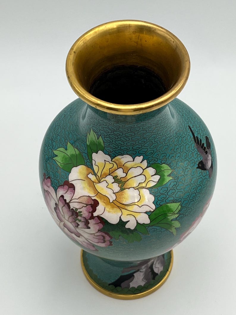 Puede incluir: Un jarr&oacute;n turquesa con borde y base dorados. El jarr&oacute;n presenta un dise&ntilde;o floral con flores amarillas, rosas y blancas, junto con un p&aacute;jaro negro. El jarr&oacute;n tiene forma redonda y un patr&oacute;n detallado.