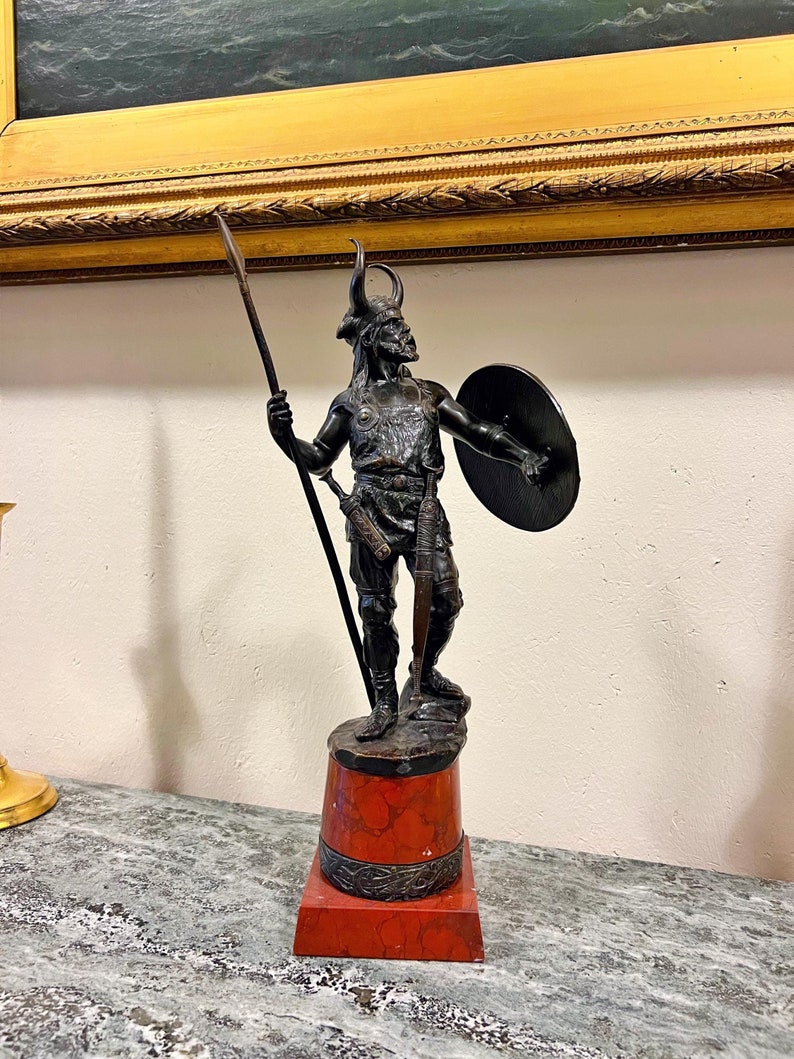 May include: A bronze sculpture of a warrior holding a spear and shield, standing on a red marble base.