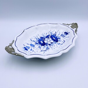 Puede incluir: Plato ovalado blanco con diseño floral azul y asas plateadas. El plato tiene un borde festoneado y un ribete azul. El diseño floral presenta flores y hojas azules. Una pieza decorativa para servir alimentos o exhibir objetos.