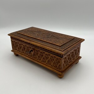May include: A rectangular wooden box with intricate carved designs. The box is a warm brown color and features a detailed pattern on the lid and sides. A small keyhole is visible on the front, suggesting it can be locked. The box rests on four small feet.