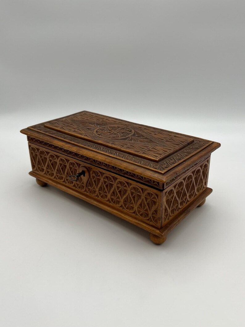 May include: A rectangular wooden box with intricate carved designs. The box is a warm brown color and features a detailed pattern on the lid and sides. A small keyhole is visible on the front, suggesting it can be locked. The box rests on four small feet.