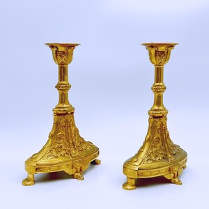 May include: Two ornate, gold-colored candle holders. Each has a detailed, floral design on the base and stem, with a small, round platform at the top for holding a candle. The candle holders are symmetrical and stand on small feet.