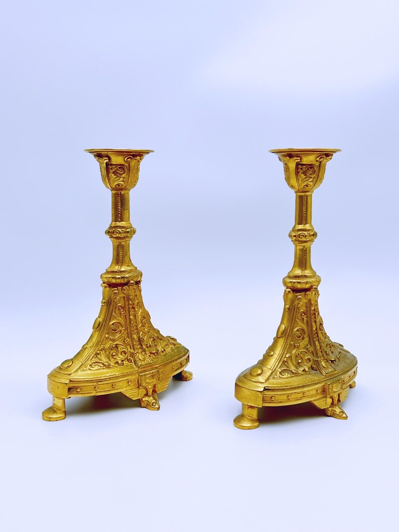 May include: Two ornate, gold-colored candle holders. Each has a detailed, floral design on the base and stem, with a small, round platform at the top for holding a candle. The candle holders are symmetrical and stand on small feet.