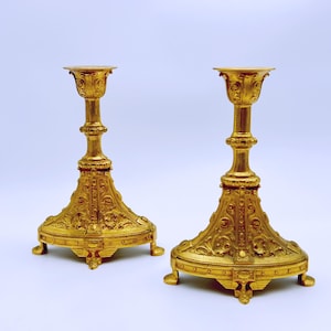 May include: Two ornate, gold-colored candle holders. Each has a wide, detailed base with a decorative pattern, a slender, textured stem, and a cup-shaped top. The candle holders are made of metal and are on a white background.