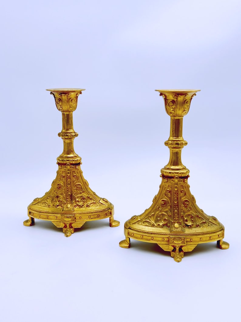 May include: Two ornate, gold-colored candle holders. Each has a wide, detailed base with a decorative pattern, a slender, textured stem, and a cup-shaped top. The candle holders are made of metal and are on a white background.