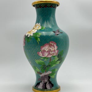 Puede incluir: Un jarr&oacute;n de cloisonn&eacute; turquesa con borde y base dorados. El jarr&oacute;n presenta dise&ntilde;os florales, incluyendo peon&iacute;as rosas y blancas, hojas verdes y un peque&ntilde;o p&aacute;jaro. El fondo tiene un patr&oacute;n texturizado.