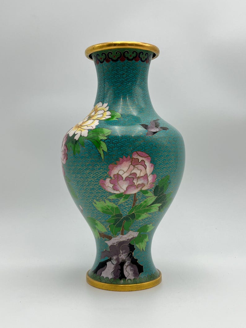 Puede incluir: Un jarr&oacute;n de cloisonn&eacute; turquesa con borde y base dorados. El jarr&oacute;n presenta dise&ntilde;os florales, incluyendo peon&iacute;as rosas y blancas, hojas verdes y un peque&ntilde;o p&aacute;jaro. El fondo tiene un patr&oacute;n texturizado.