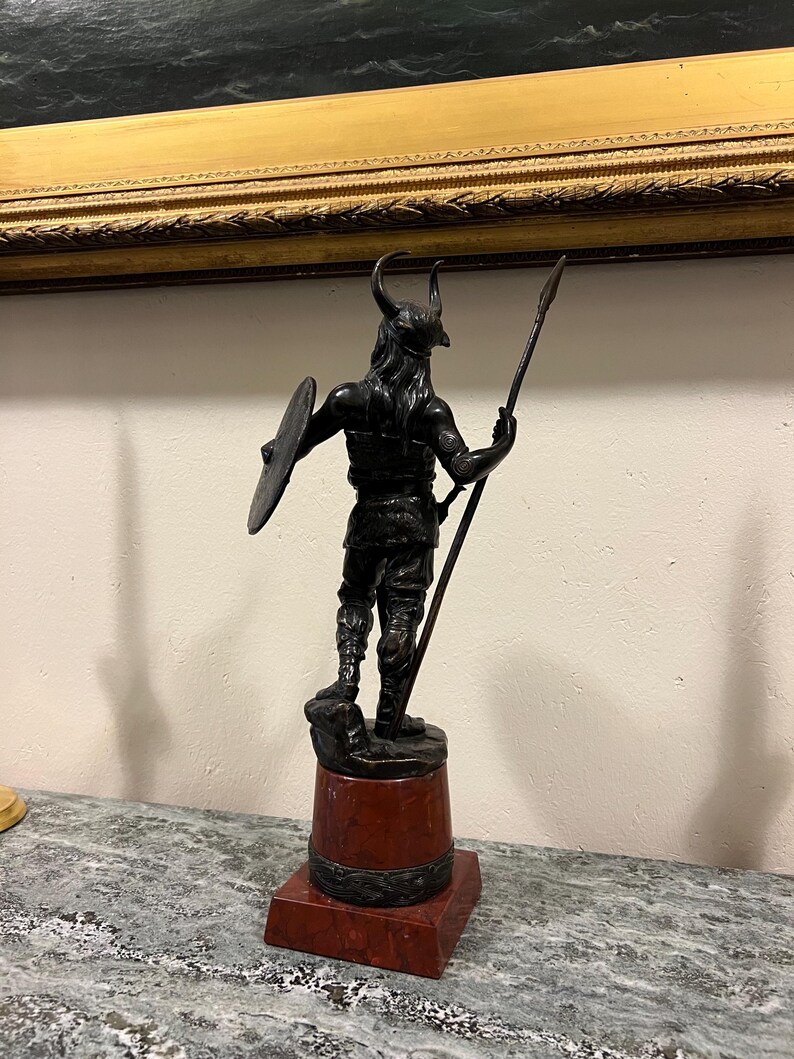 May include: A bronze sculpture of a Viking warrior holding a spear and shield, standing on a red marble base.