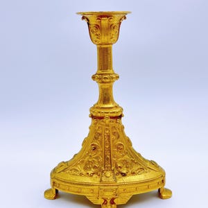 May include: Ornate gold-colored candlestick with a wide, detailed base and a slender, decorated stem. The top features a flared cup shape. The candlestick has a classical design with intricate patterns and three small feet.