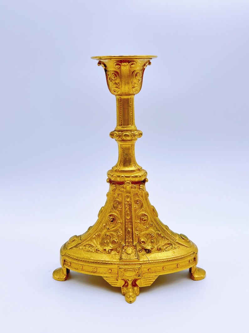 May include: Ornate gold-colored candlestick with a wide, detailed base and a slender, decorated stem. The top features a flared cup shape. The candlestick has a classical design with intricate patterns and three small feet.