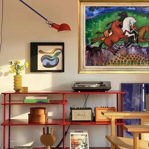 May include: A red metal shelving unit displays a turntable, speakers, and decorative items. A framed painting of horses hangs above the unit. A red lamp extends from the wall. A yellow vase holds flowers. A wooden sculpture and books are also visible.