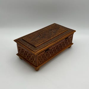 May include: A rectangular wooden box with intricate carved designs. The box is a warm brown color and features detailed patterns on the lid and sides. It rests on small, rounded feet.