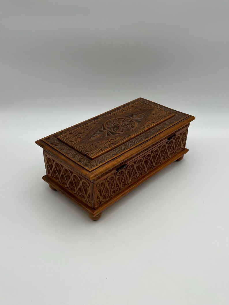 May include: A rectangular wooden box with intricate carved designs. The box is a warm brown color and features detailed patterns on the lid and sides. It rests on small, rounded feet.