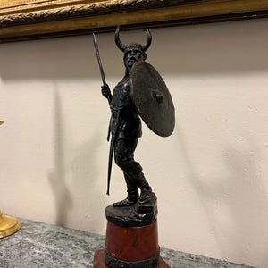 May include: A bronze sculpture of a Viking warrior holding a spear and shield. The sculpture is mounted on a red marble base.